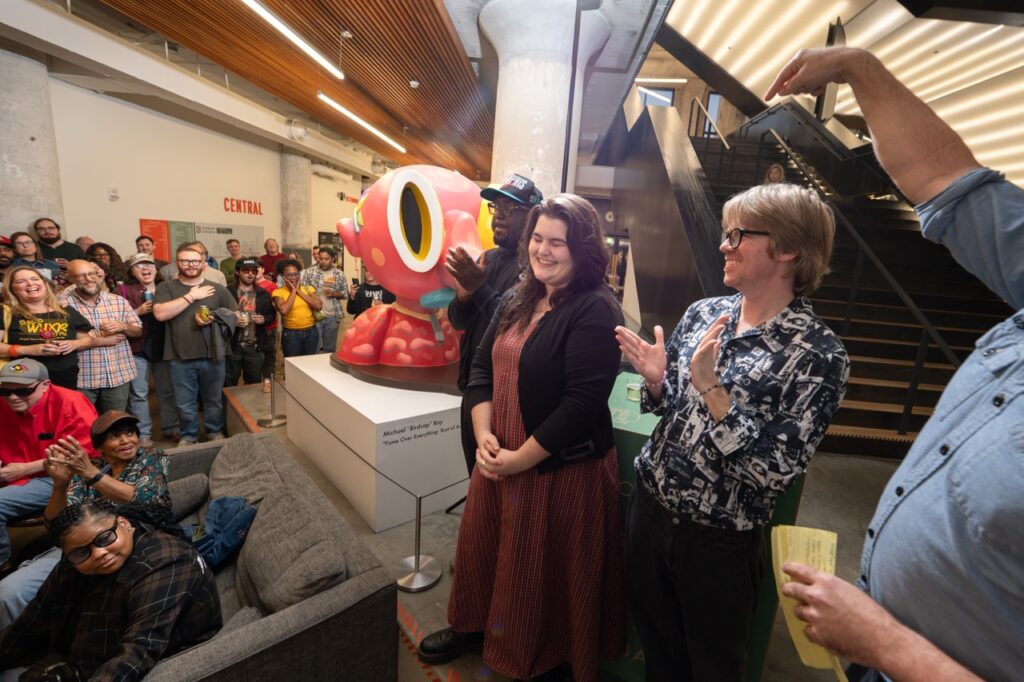 Three people stand by the pink sculpture, smiling, as the crowd watches.