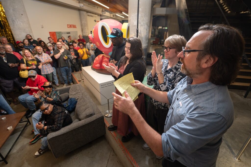 Speakers applaud while a man reads from a yellow sheet of paper.