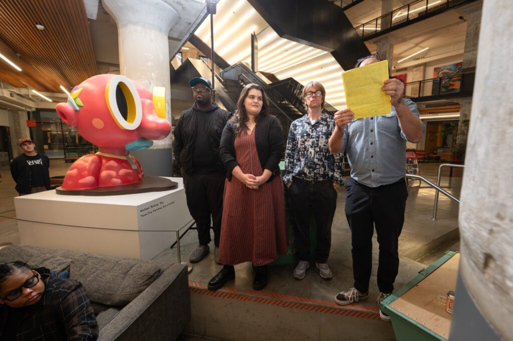 Four people stand at the front by the pink sculpture during the program.