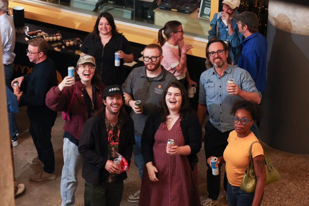 Overhead view of party guests smiling and raising cans toward the camera in a toast.