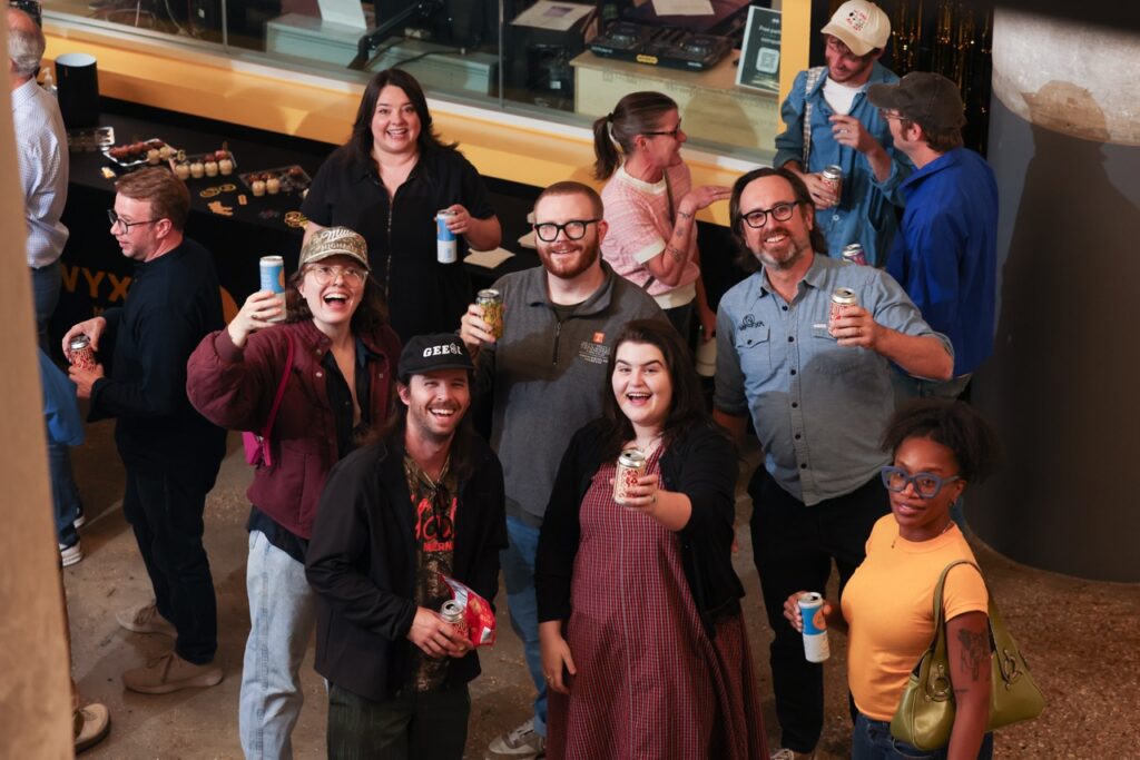 Overhead view of party guests smiling and raising cans toward the camera in a toast.