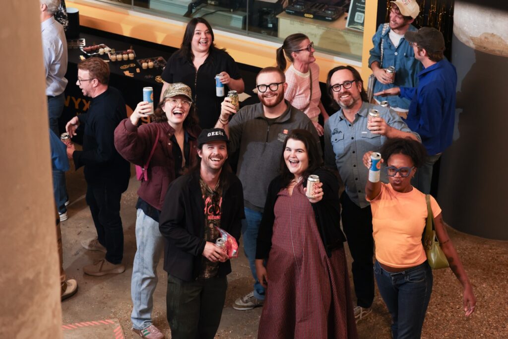 Overhead view of party guests smiling and raising cans toward the camera in a toast.