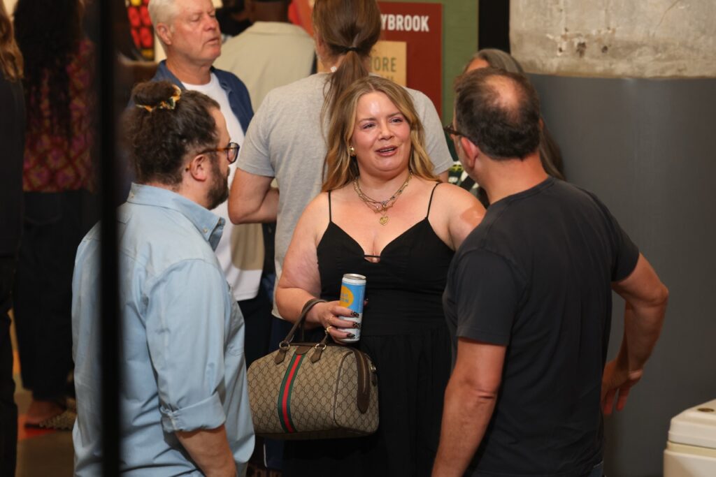 A woman in a black dress holding a can chats with guests in a crowded room.