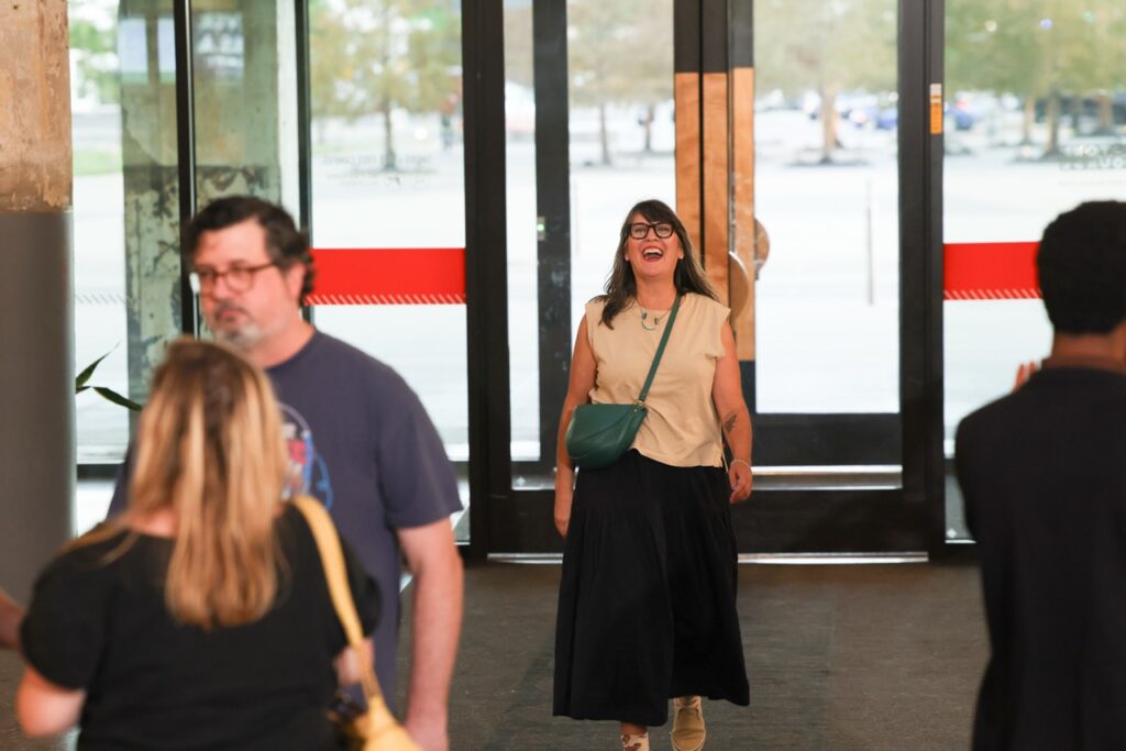A smiling woman in a cream top and long black skirt walks through glass doors into the party.