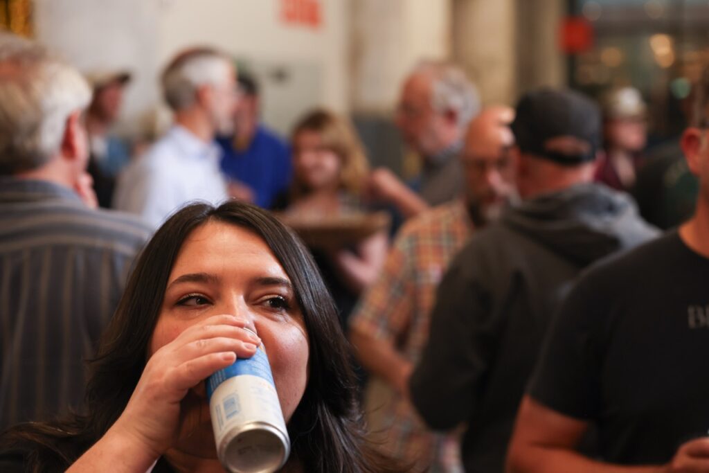 A woman tips back a can of beer in the middle of a busy crowd.