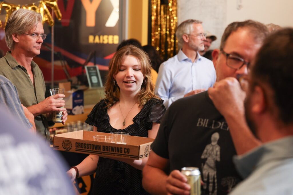 A woman in black carries a box of drinks through the crowd, with guests holding cans around her.