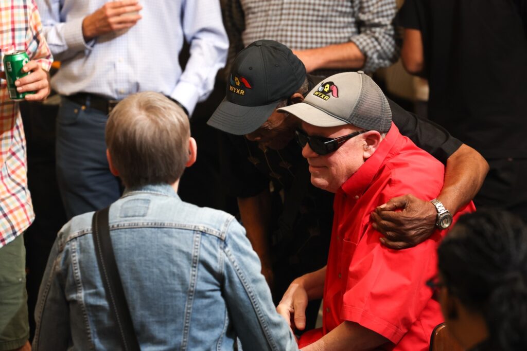 A man in a red shirt and sunglasses is hugged by a friend while guests in caps stand nearby.