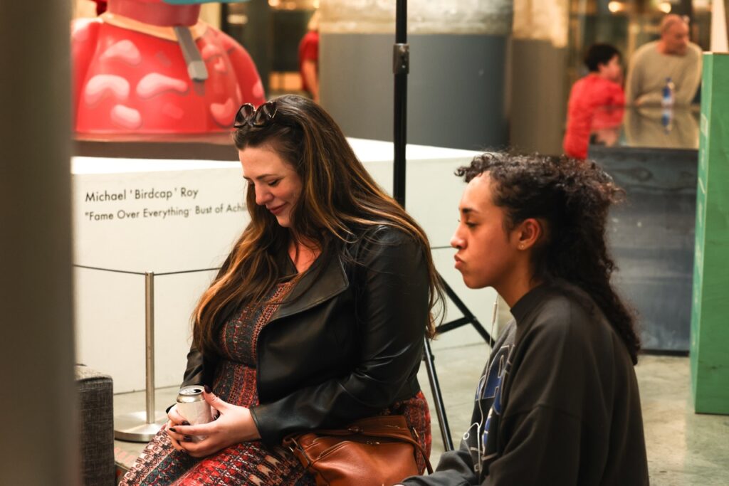 Two women sit together near a gallery display, one smiling down at the can in her hands.