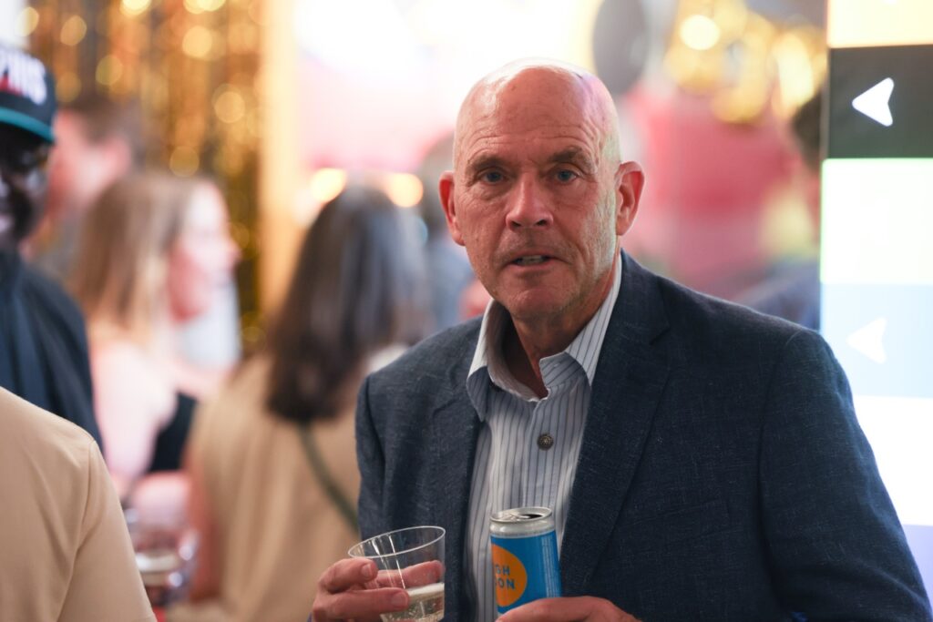 A man in a navy blazer holds a cup and a can while looking toward the camera at the party.