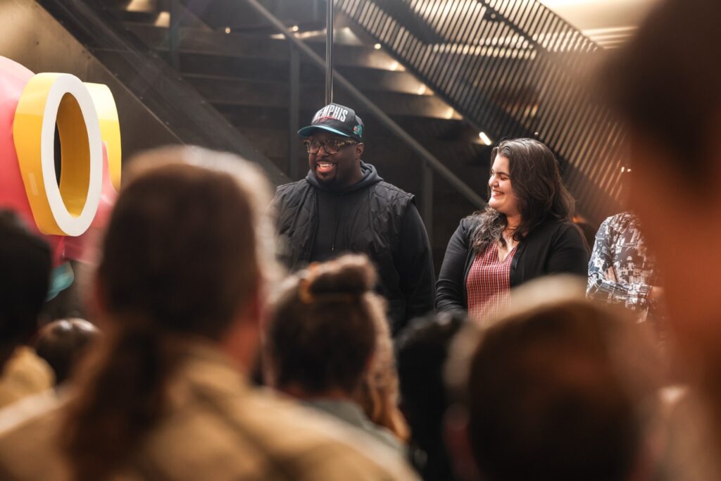 A man in a black cap and a woman in a red dress stand in front of the crowd before the toast.