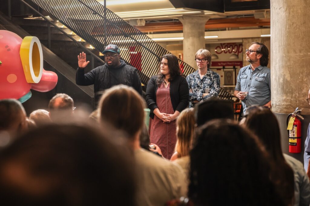 A man in a black cap raises a hand to the crowd as a woman and two men stand beside him.