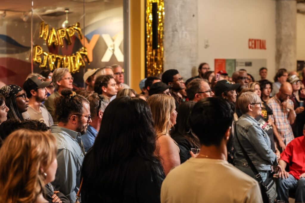A packed crowd of guests fills the atrium under the gold HAPPY BIRTHDAY balloons.