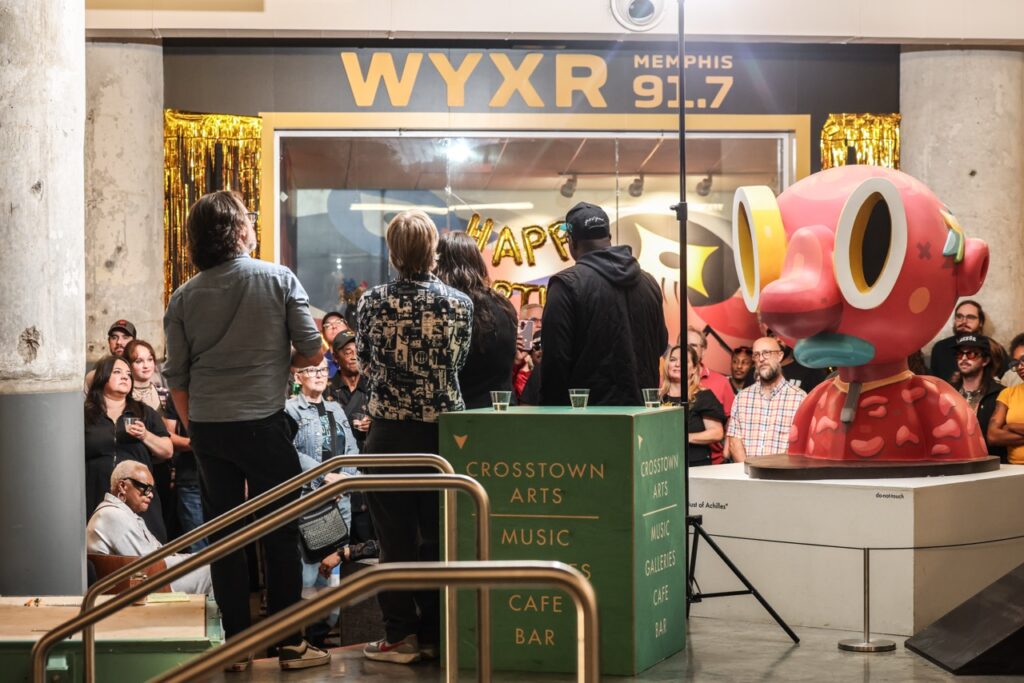Guests face the WYXR studio window, decorated with gold balloons, with a large pink sculpture to the right.