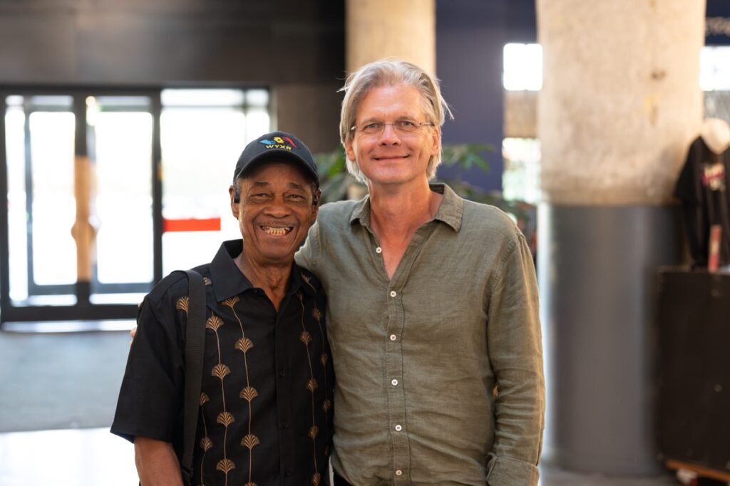 Two men stand close together smiling for a photo, one in a patterned dark shirt and one in a green button-up.