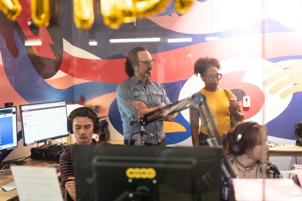 Through the studio window, a DJ in headphones sits at the board while two visitors stand behind with a colorful mural behind them.
