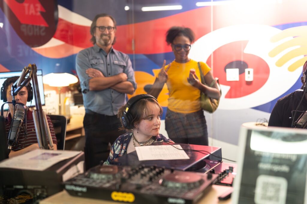 Through the studio window, a DJ in headphones sits at the board while two visitors stand behind with a colorful mural behind them.