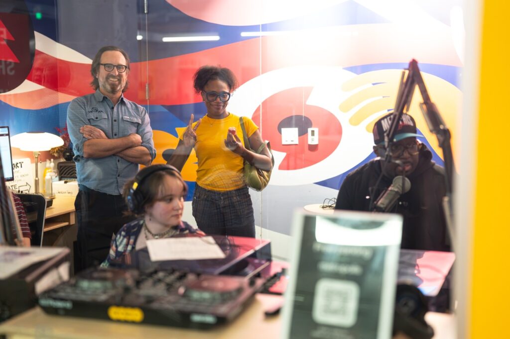 Visitors stand in the WYXR studio behind a DJ in headphones, seen through the window.