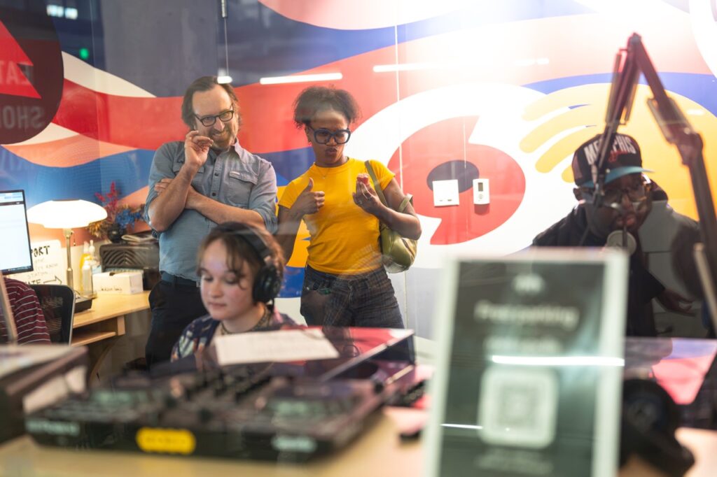 Visitors stand in the WYXR studio behind a DJ in headphones, seen through the window.