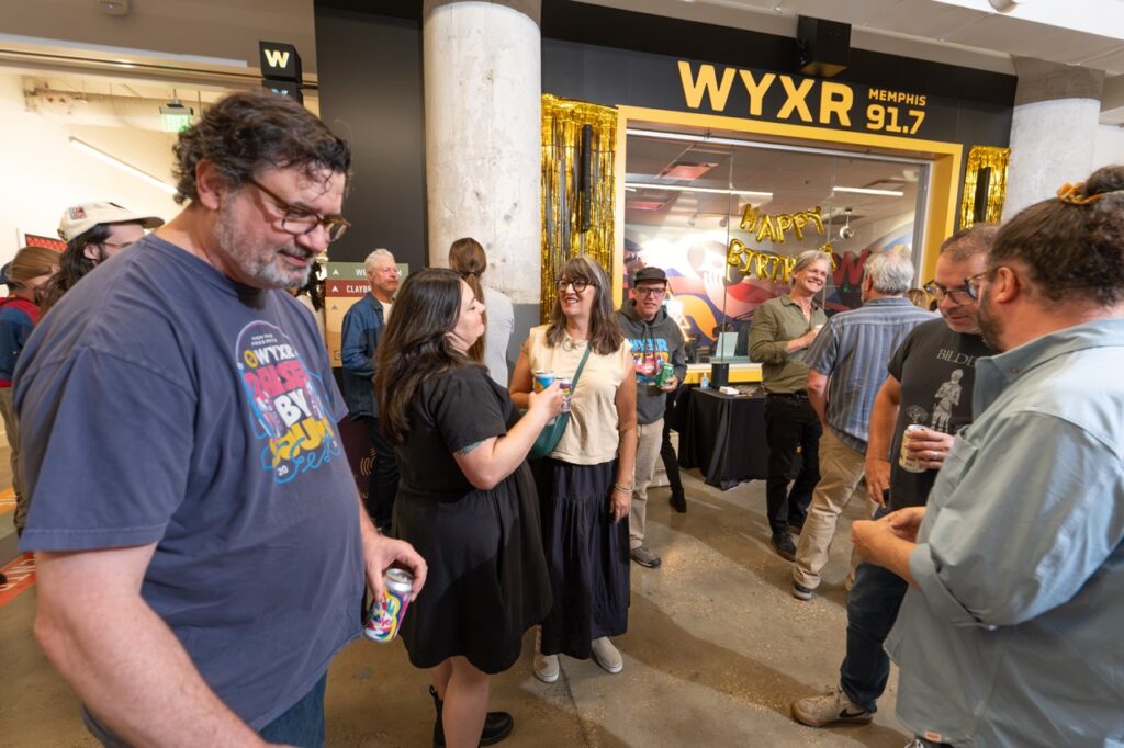 A man in a blue WYXR T-shirt stands in the foreground as guests gather near the lit WYXR 91.7 studio window.