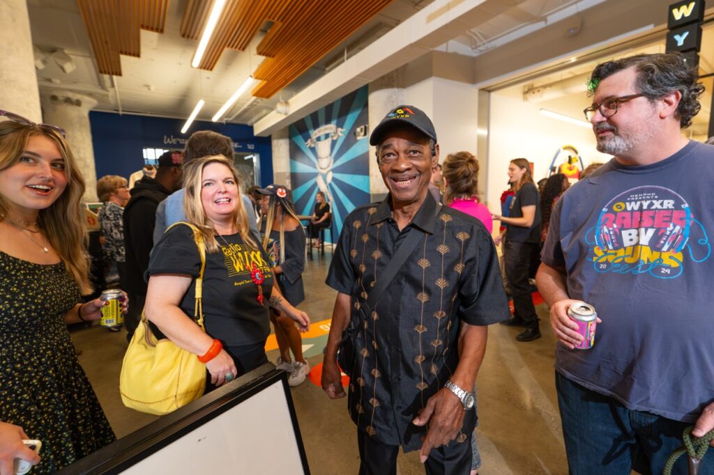 Guests mingle in the atrium, with a man in a cap and WYXR shirt talking to a friend.