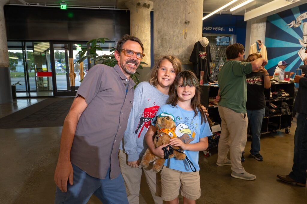 A man poses with two kids in colorful T-shirts who are holding stuffed animals.
