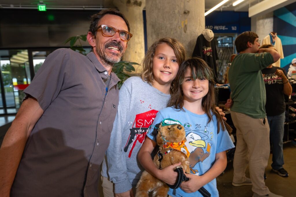 A man poses with two kids in colorful T-shirts who are holding stuffed animals.