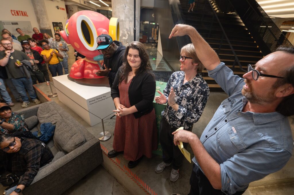 Three people stand by the pink sculpture as one man points up with a raised arm.