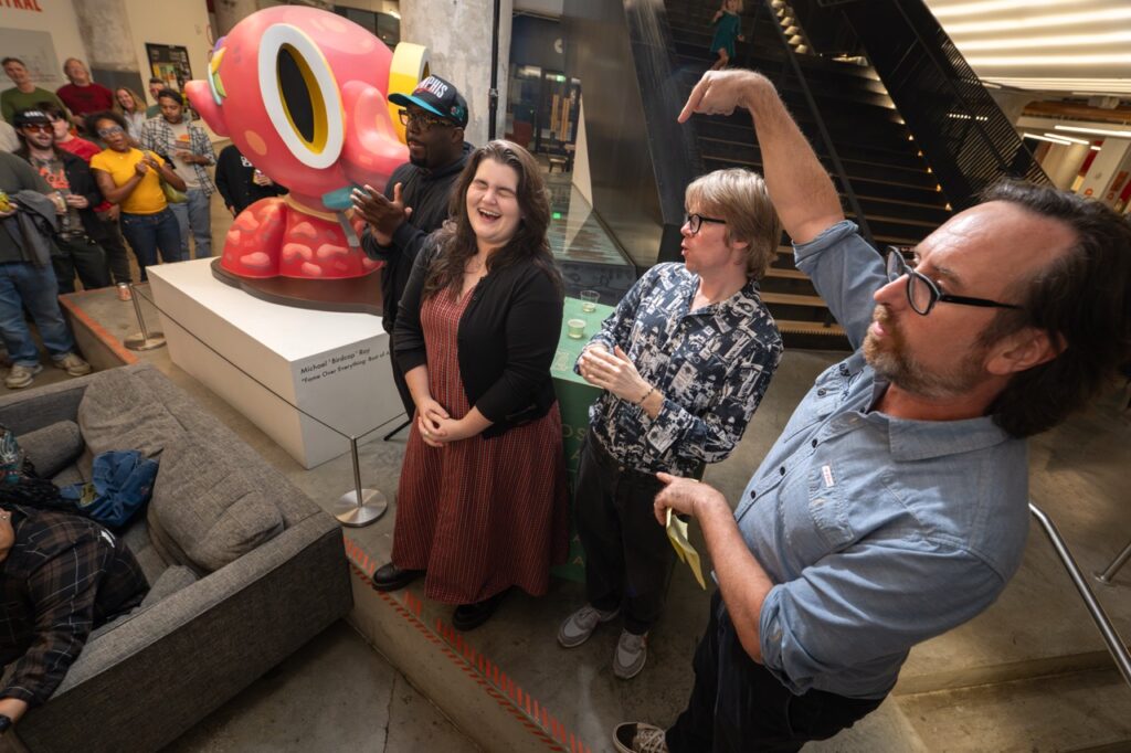 Three people stand by the pink sculpture as one man points up with a raised arm.