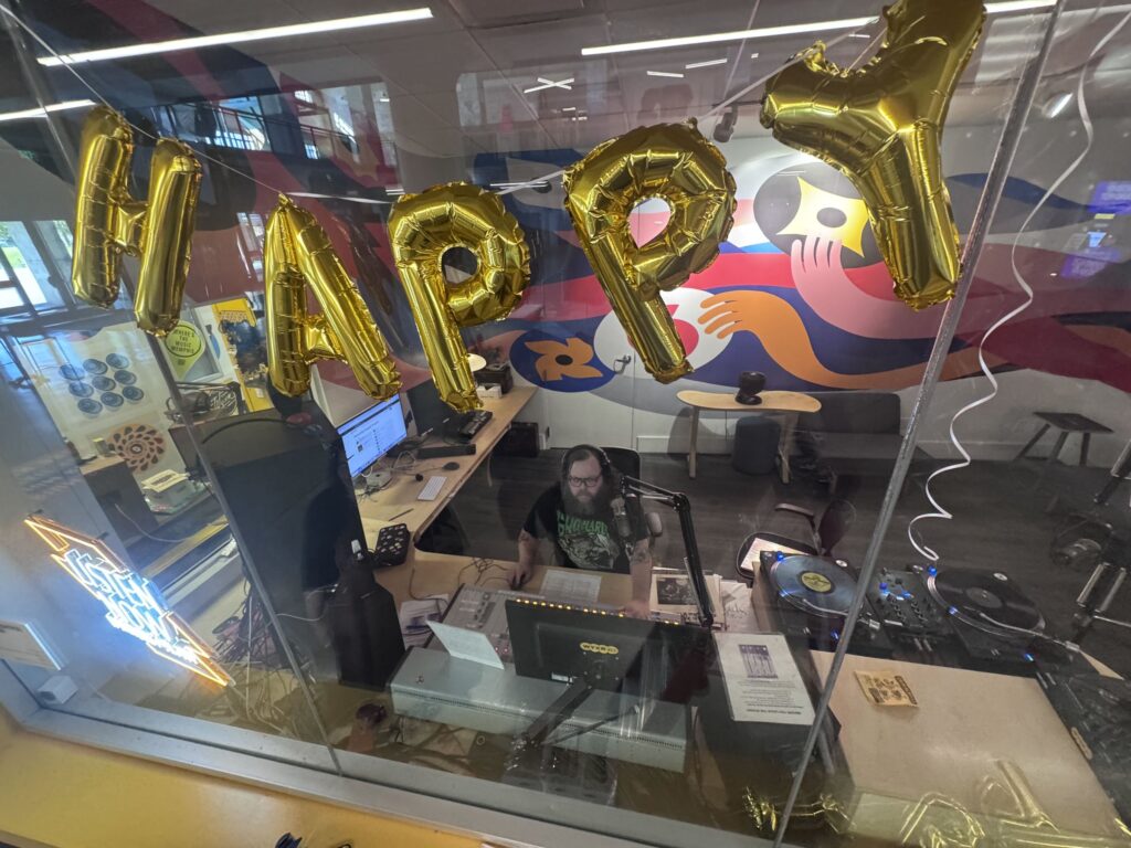 View through a WYXR studio window decorated with gold foil HAPPY letter balloons, showing a DJ wearing headphones at the mixing console with turntables to the right and a colorful mural behind.