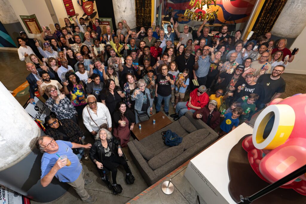 Overhead view of a large crowd of WYXR staff, volunteers, DJs and friends raising drinks in a toast in a concrete-columned atrium, with gold HAPPY BIRTHDAY balloons above the studio window and colorful murals behind them.